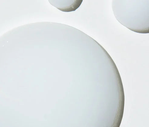 A close-up studio shot of a glossy, opaque white cosmetic droplet or gel sphere on a clean white background, with partial additional droplets visible at the top edge.