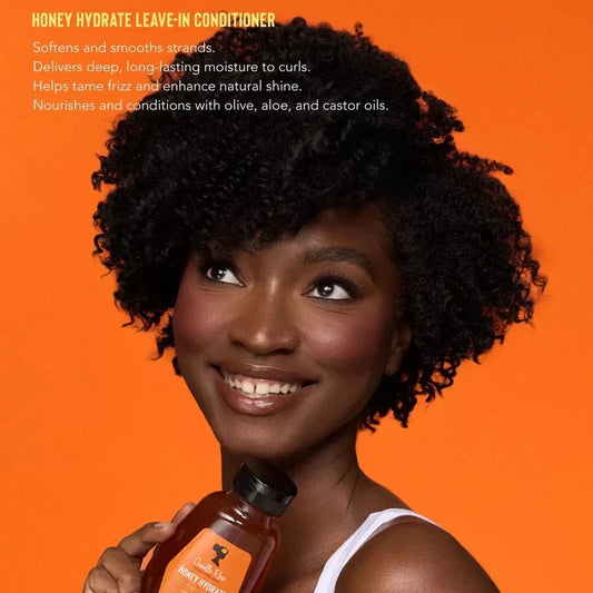 A smiling woman with voluminous curly hair holds a bottle of Honey Hydrate Leave-In Conditioner against a bright orange background, with product text visible on the left side of the image.