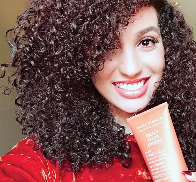 A smiling woman with voluminous dark brown curly hair holds a tube of Miker O'Callaghan Daily Hair Dress conditioner. The background is a soft, warm beige tone.