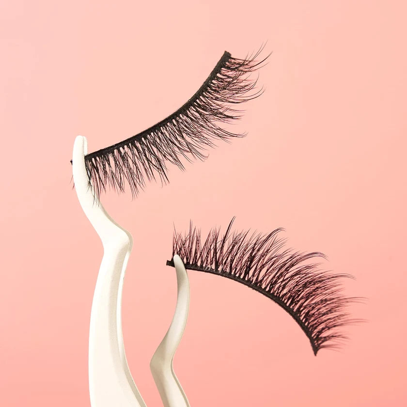 Two pairs of black false eyelashes displayed on curved white stands against a soft pink background.