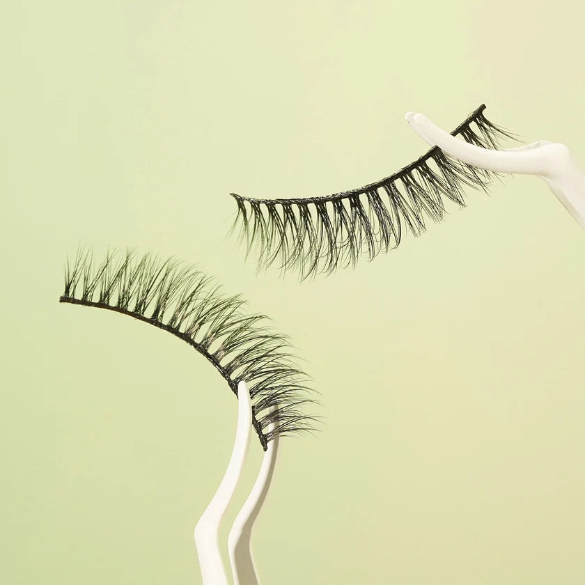 A pair of black false eyelashes displayed on white curved stands against a pale yellow-green background.