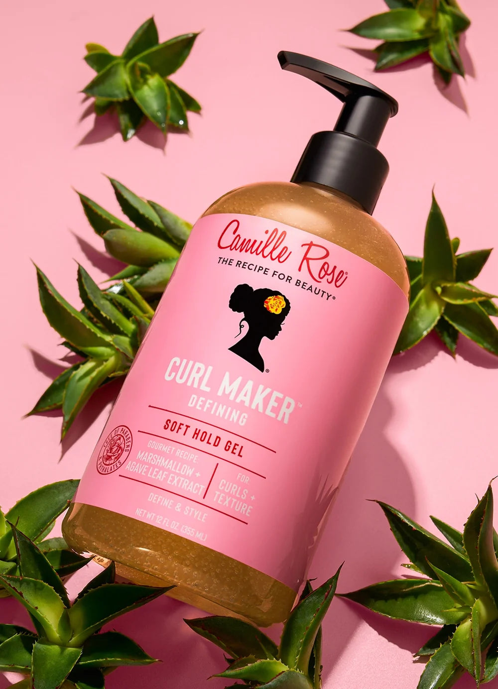 Camille Rose Curl Maker Soft Hold Gel bottle on a pink background with green leaves.
