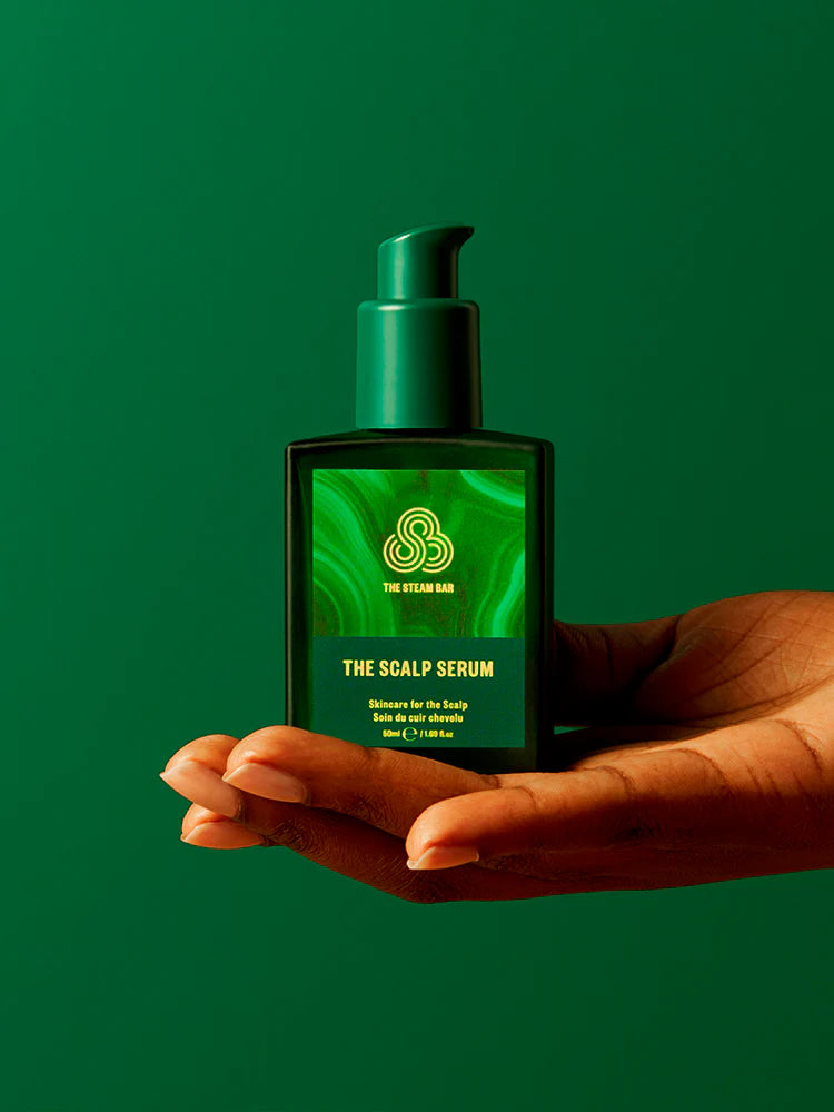Hand holding a bottle of 'The Scalp Serum' against a green background