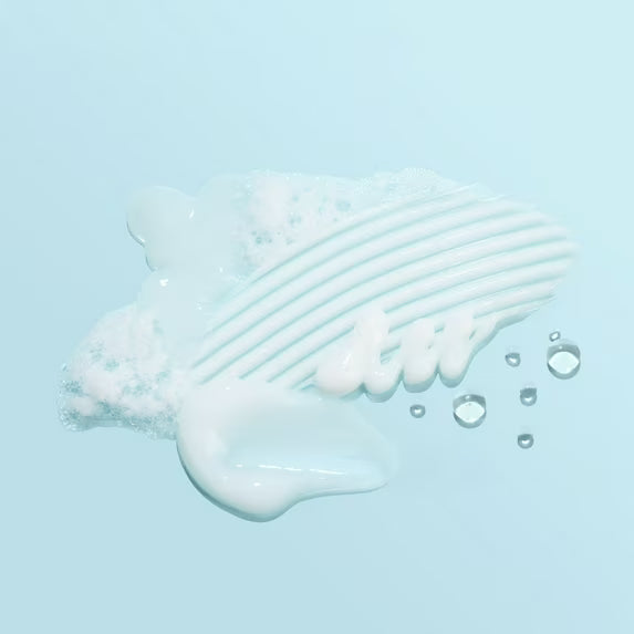 A close-up of a white foaming cleanser or soap being spread across a ridged surface, with small bubbles floating nearby, on a light blue background.