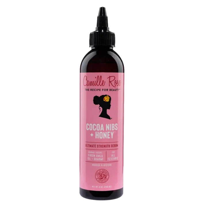 A bottle of Camille Rose Cocoa Nibs + Honey hair serum with a pink label and black cap, shown on a plain white background.