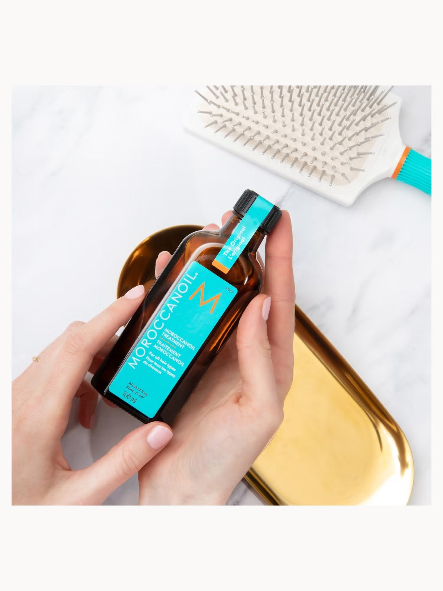 Person holding a bottle of Moroccanoil product with a hairbrush in the background