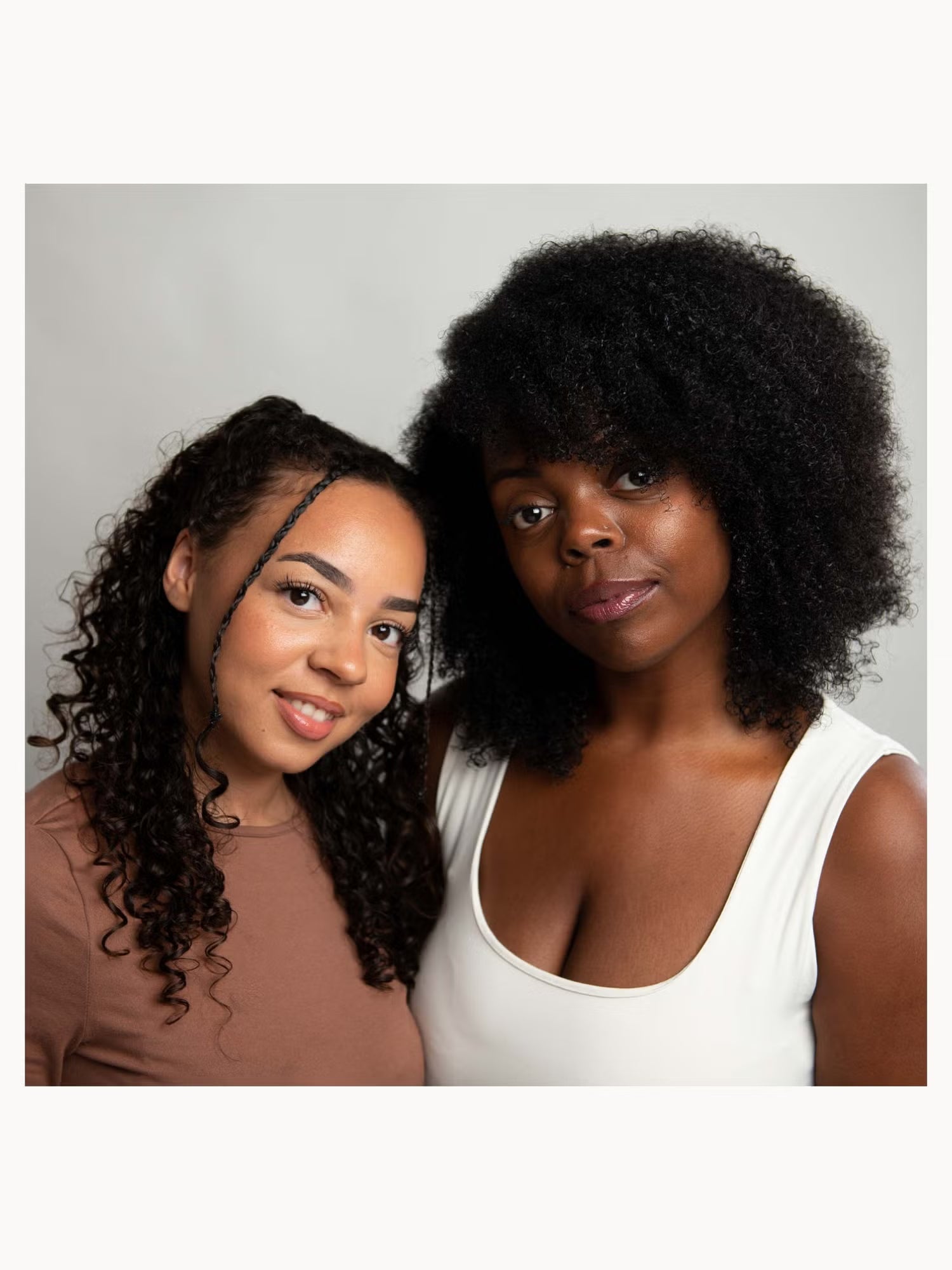 Two women posing together against a light gray studio background. The woman on the right has voluminous dark curly hair and wears a white tank top, while the woman on the left has long dark curly hair and wears a brown top.