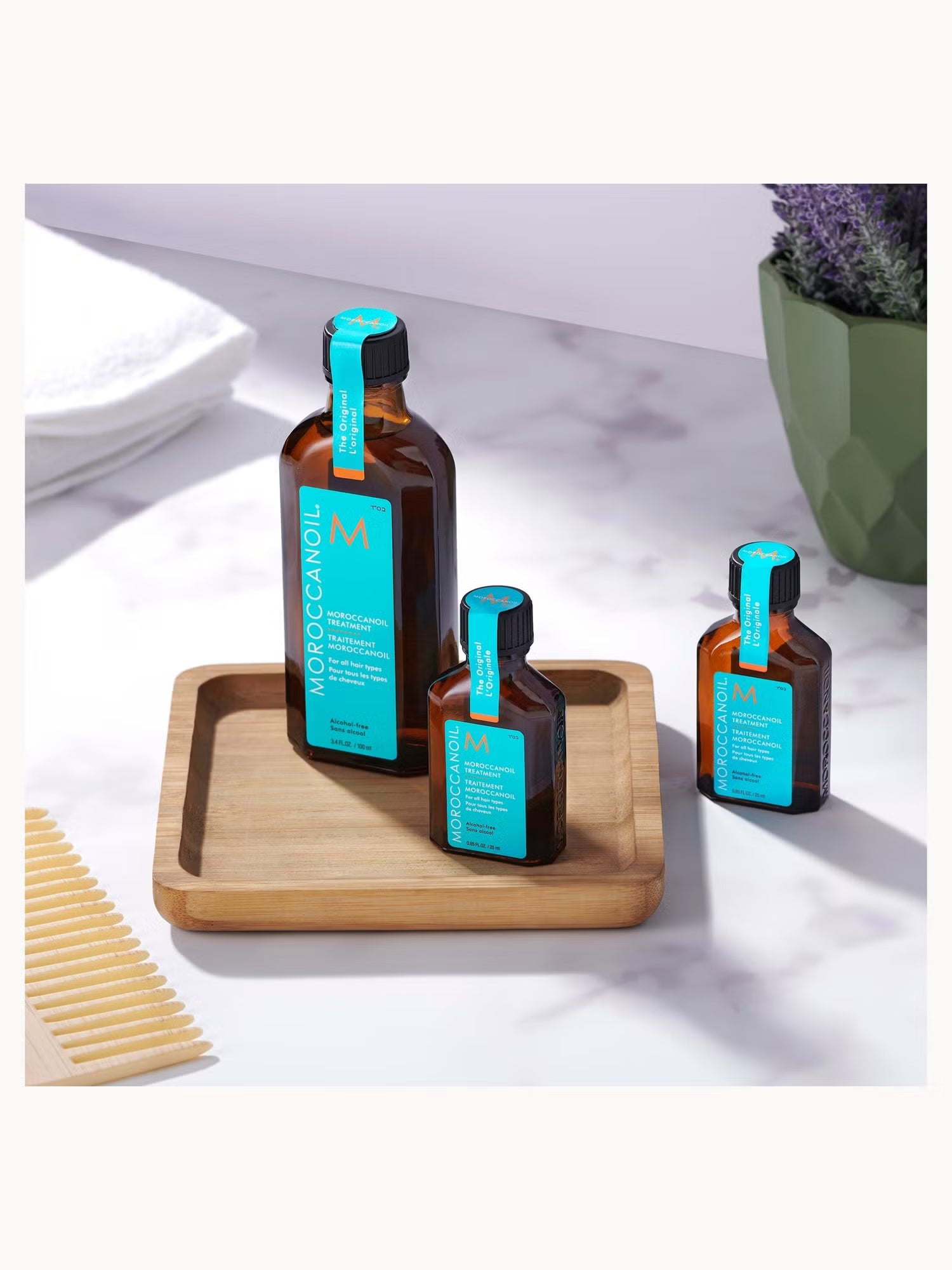 Three bottles of Moroccanoil product on a wooden tray with a comb and plant in the background.