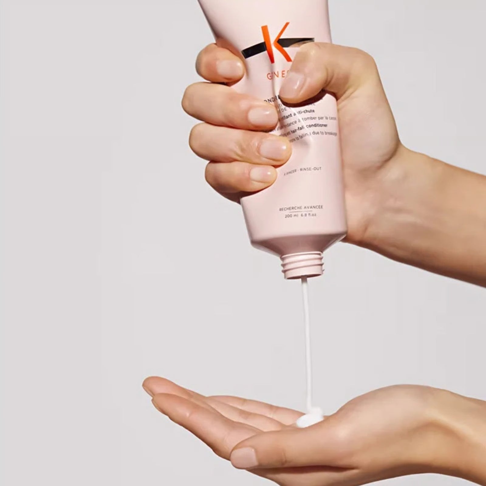 Hand holding a pink K-ONE hair product bottle with visible brand logo, dispensing product onto another hand against a light gray background.