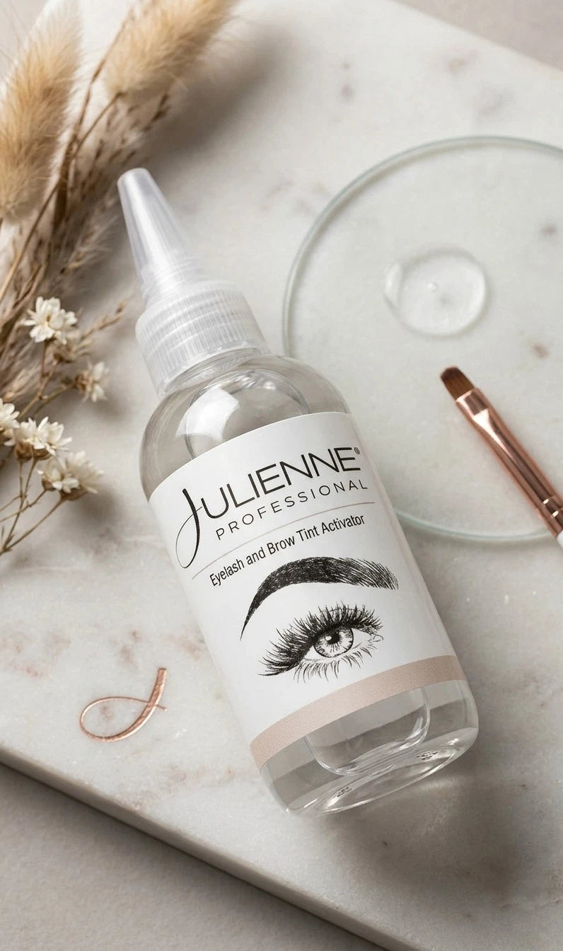 A clear plastic bottle of Julienne Professional Eyelash and Brow Tint Activator with a white label, styled on a light marble surface with cosmetic brushes and dried grass.