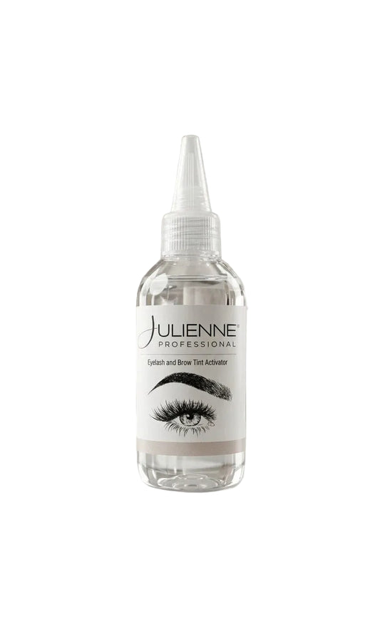 A clear bottle of Julienne Professional eyelash and brow tint activator with a white label, displayed on a white marble block in a softly lit vanity setting.