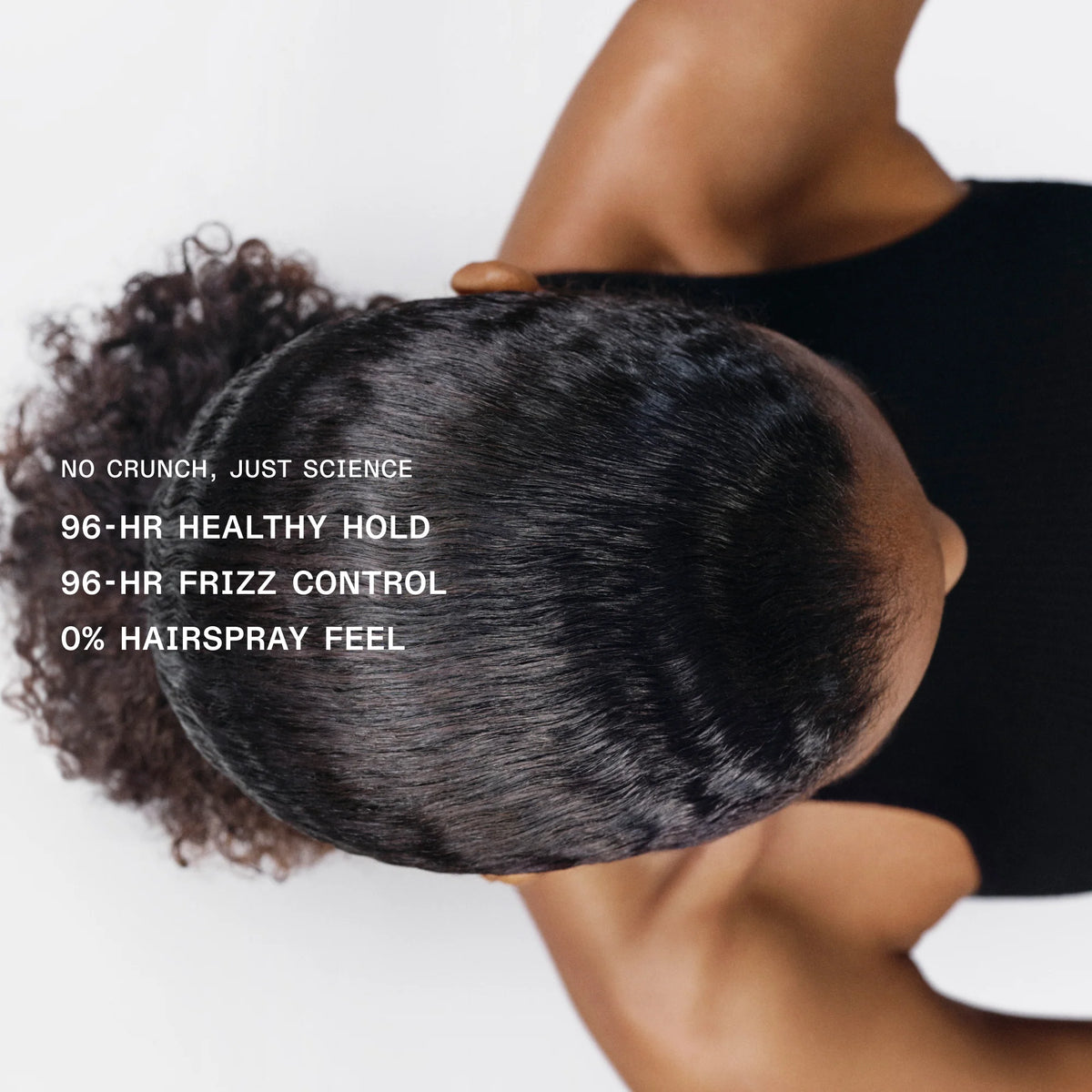 Top-down view of a person wearing a black top, showing the back of their head with dark curly hair styled in a low bun. White text on the left reads: 'NO CRUNCH, JUST SCIENCE 96-HR HEALTHY HOLD 96-HR FRIZZ CONTROL 0% HAIRSPRAY FEEL'.